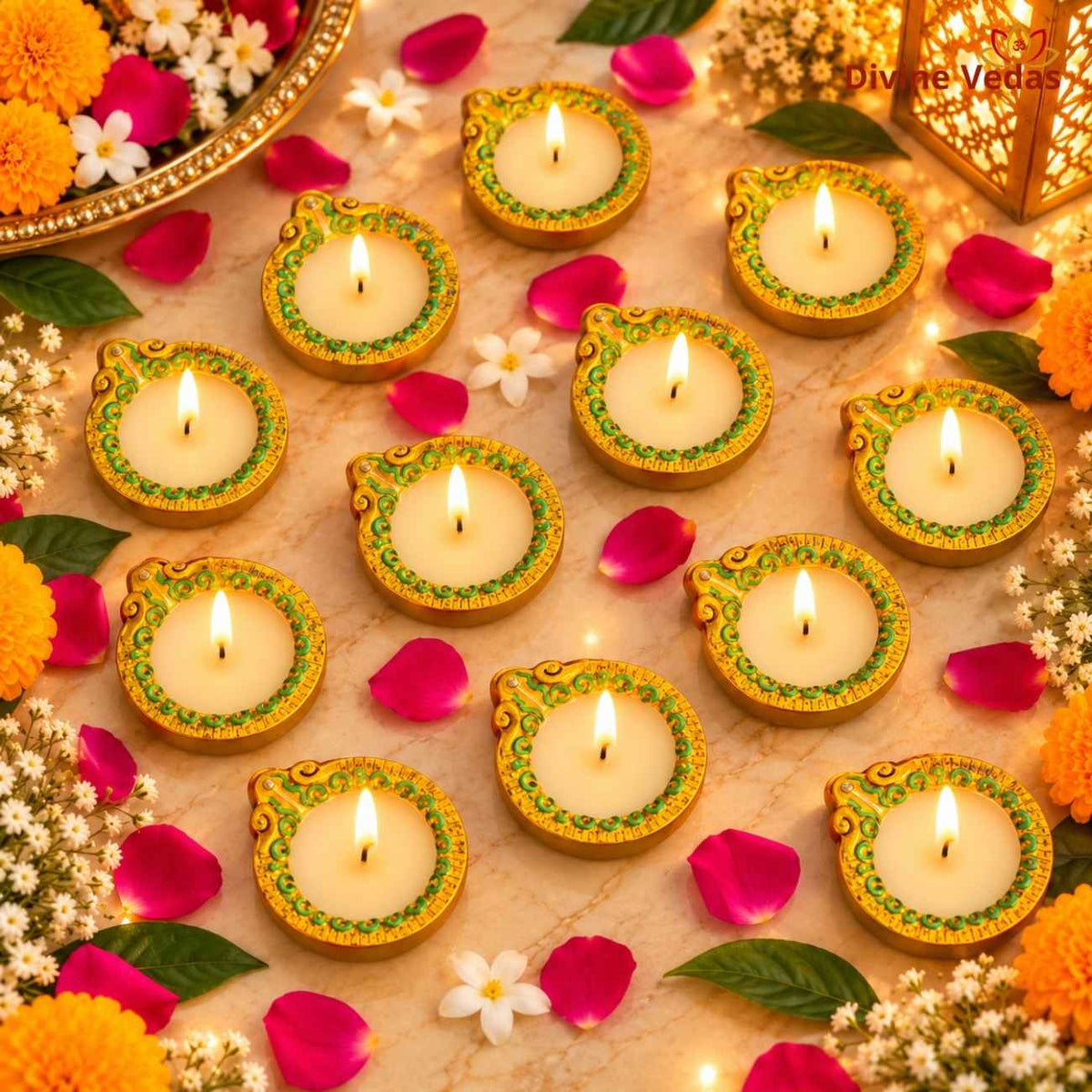 A decorative flat lay of multiple lit diya candles arranged on a marble surface with flowers, petals, green leaves, and warm string lights. The candles have cream-colored wax and gold-toned rims with green floral detailing.